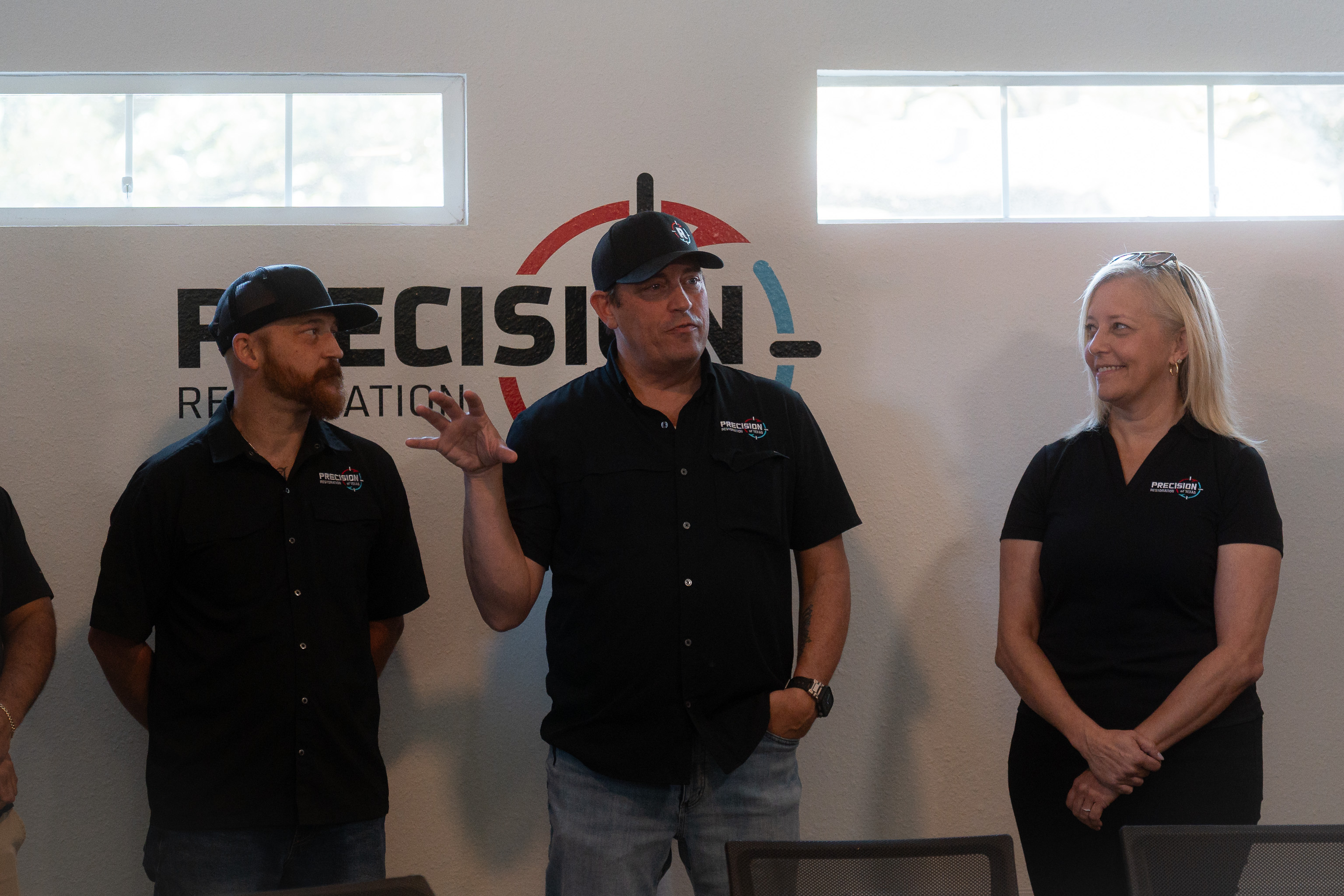 Three people wearing Precision Restoration shirts standing and talking in front of a company logo wall.