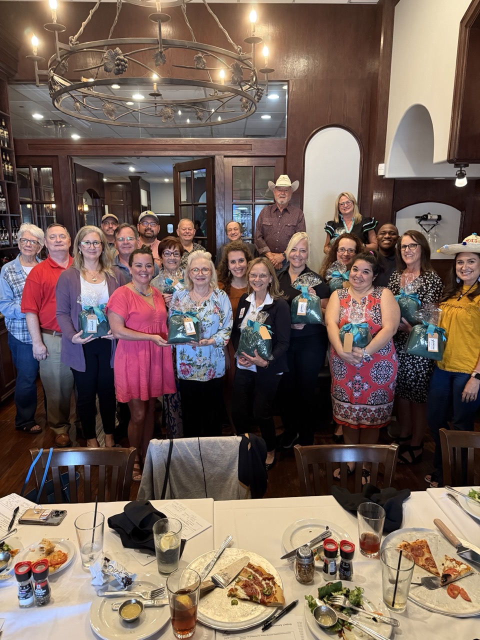 Group of people posing in a dining room holding wrapped gifts with tables of food in front.