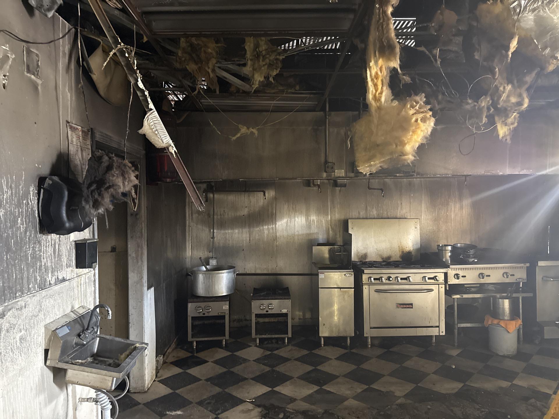 Fire-damaged commercial kitchen with soot-covered walls, exposed ceiling insulation, and dirty checkered floor.
