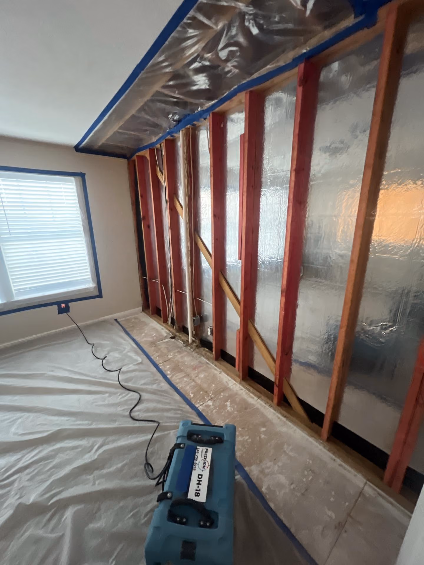 Room wall stripped to red wooden studs with reflective insulation and a blue dehumidifier on plastic-covered floor.