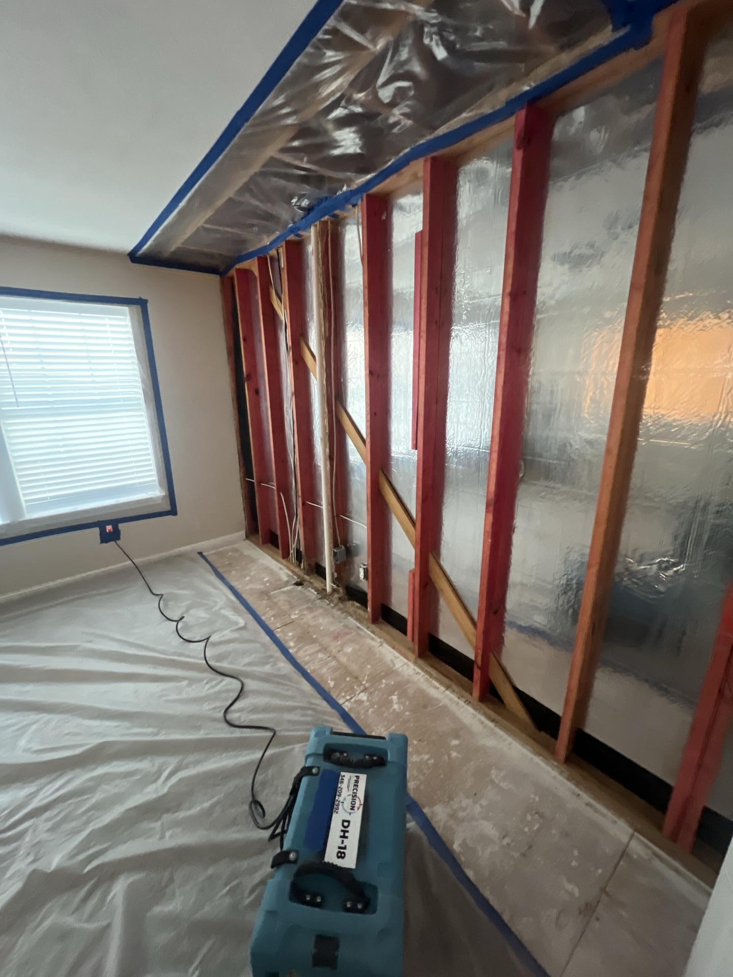 Room wall stripped to red wooden studs with reflective insulation and a blue dehumidifier on plastic-covered floor.