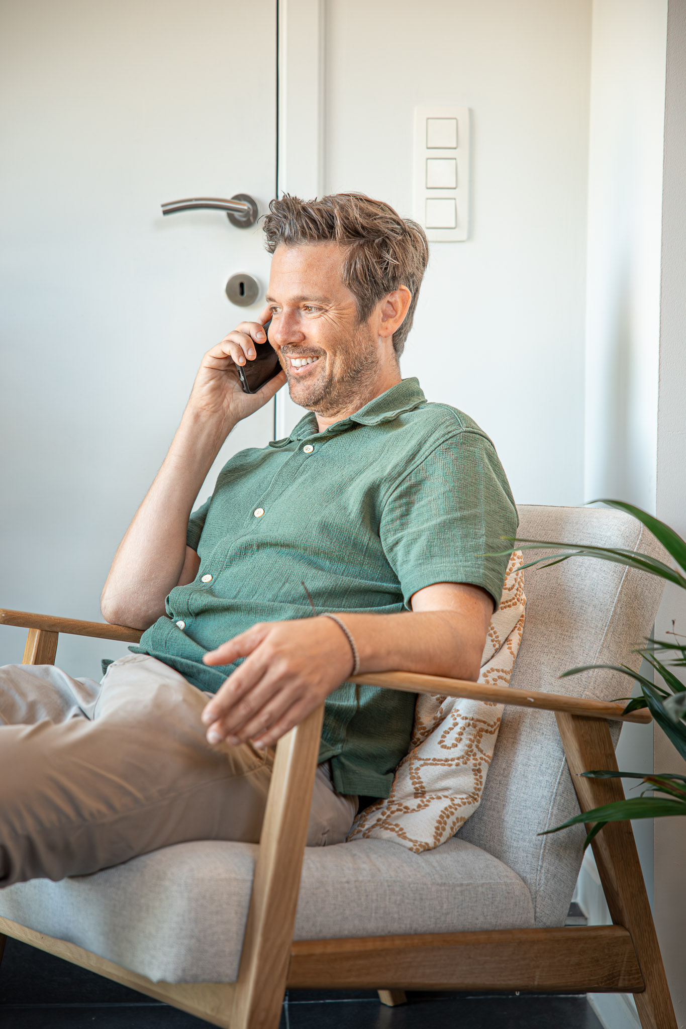 Man in een groene polo zittend in een houten stoel met een kussen, glimlachend terwijl hij telefoneert in een heldere kamer.