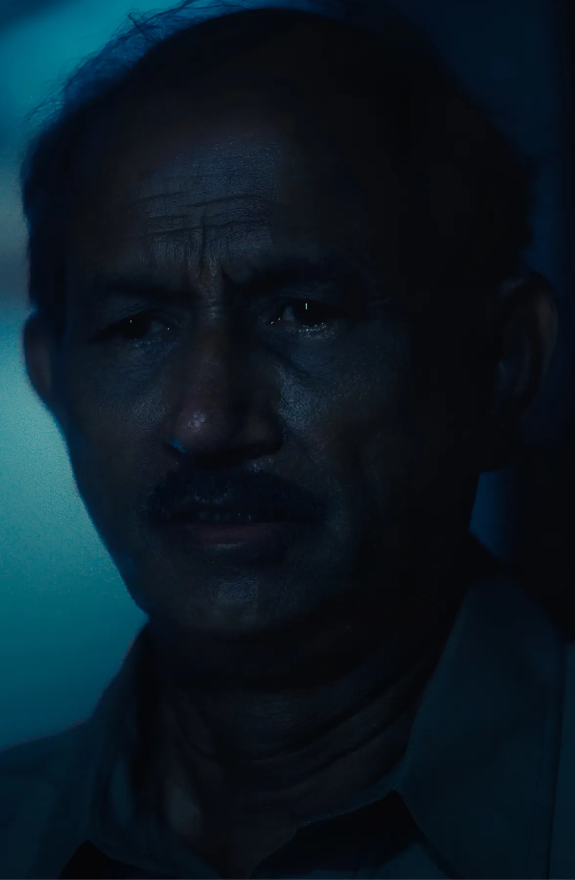 Close-up of a man with a mustache in dim blue lighting, showing a serious expression.