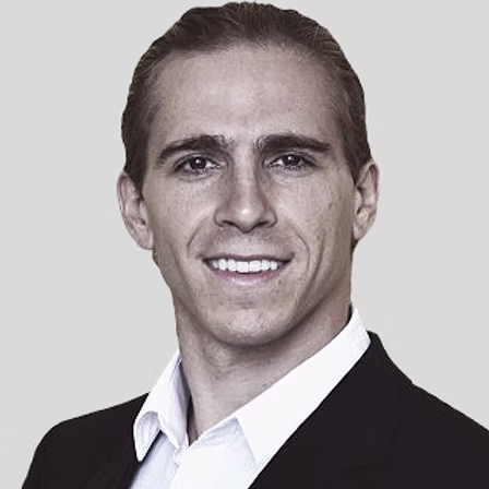 Smiling man with slicked-back hair wearing a white shirt and black jacket against a light background.