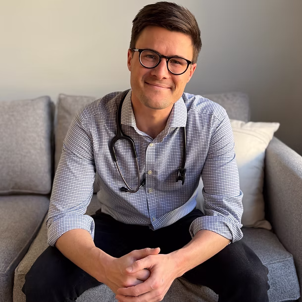 Smiling man with glasses and stethoscope sitting on gray couch with hands clasped.