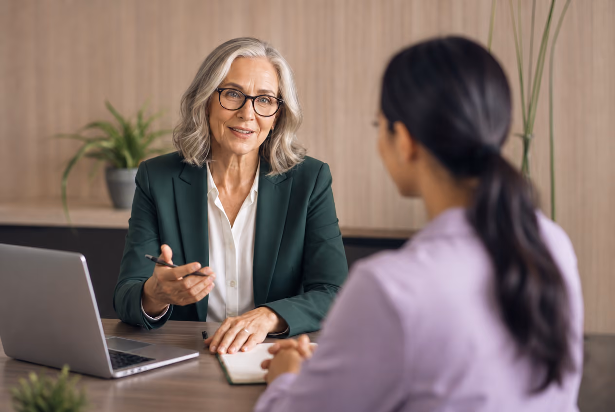 Mature woman in glasses discussing with younger woman at a desk with laptop and notebook.