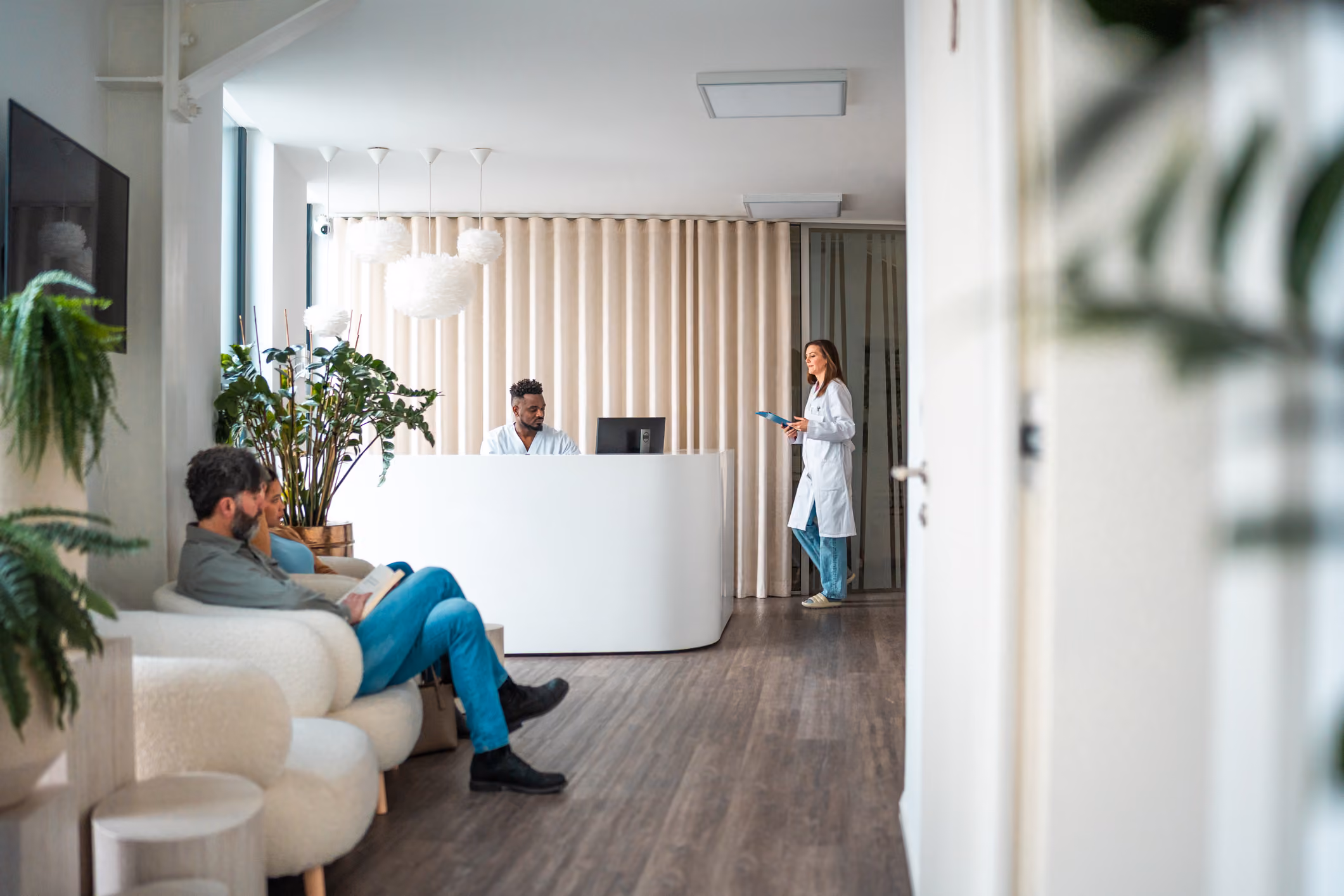 Patients seated in a medical waiting room with a receptionist and a doctor standing behind a white desk.