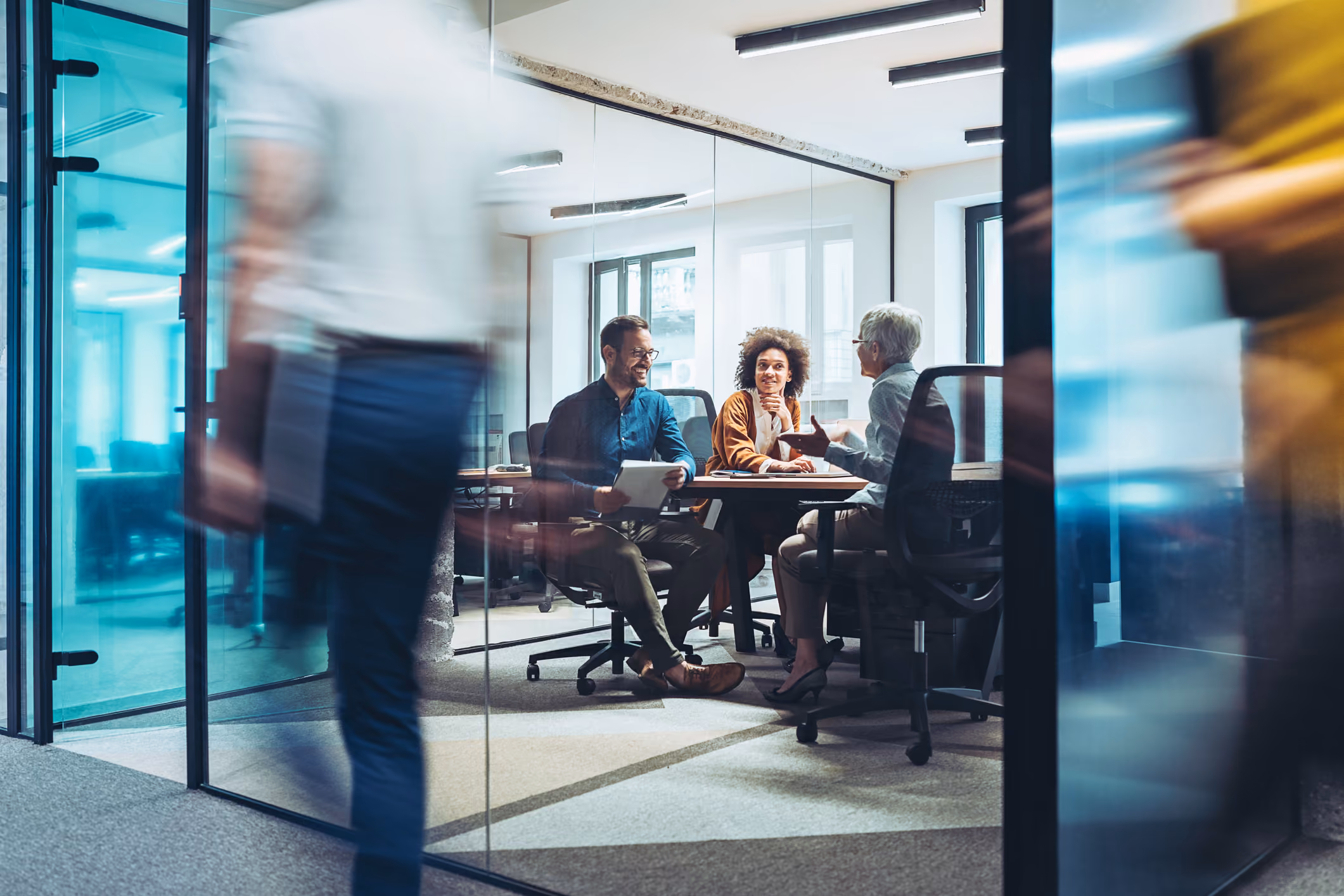 Three colleagues sitting at a table having a discussion inside a glass-walled office room.