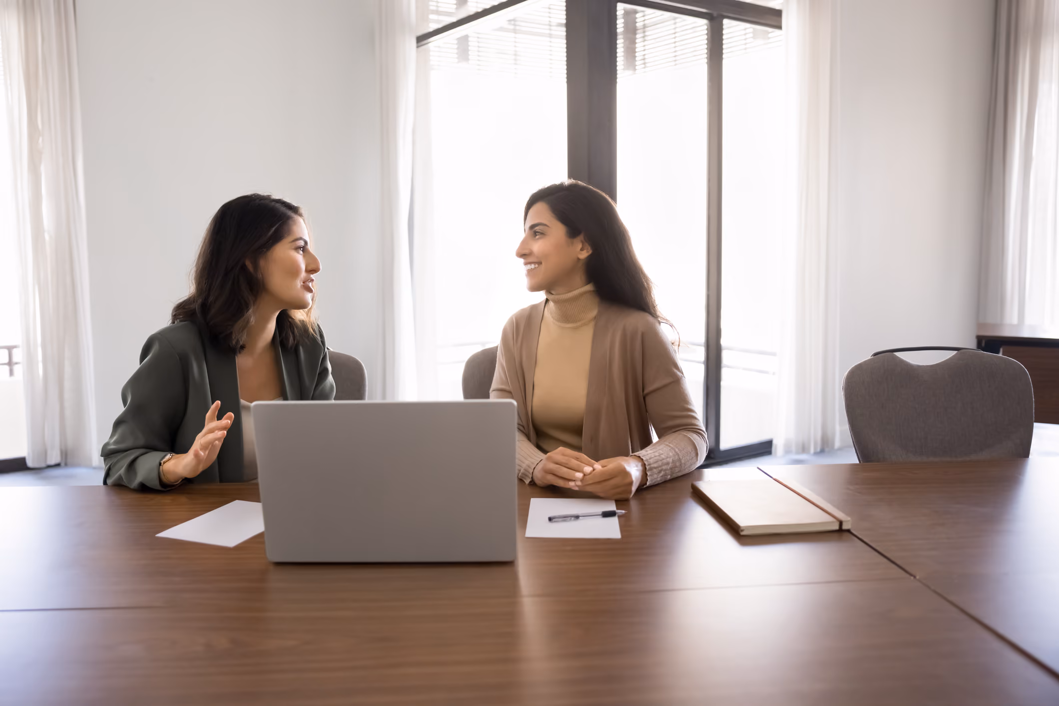Two women sitting at a conference table with laptop and papers, engaged in conversation.