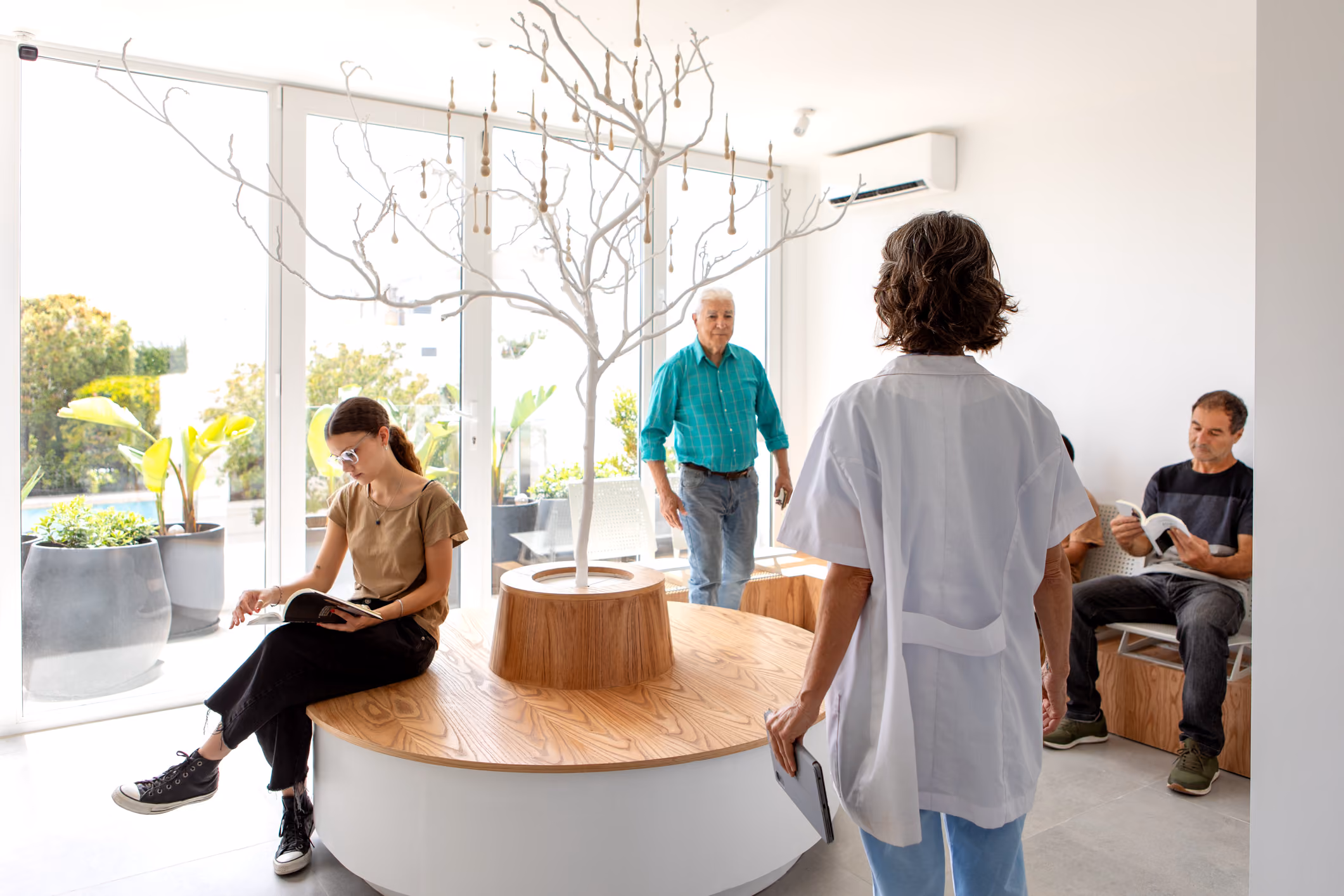 People in a bright waiting room with large windows and a decorative tree centerpiece.