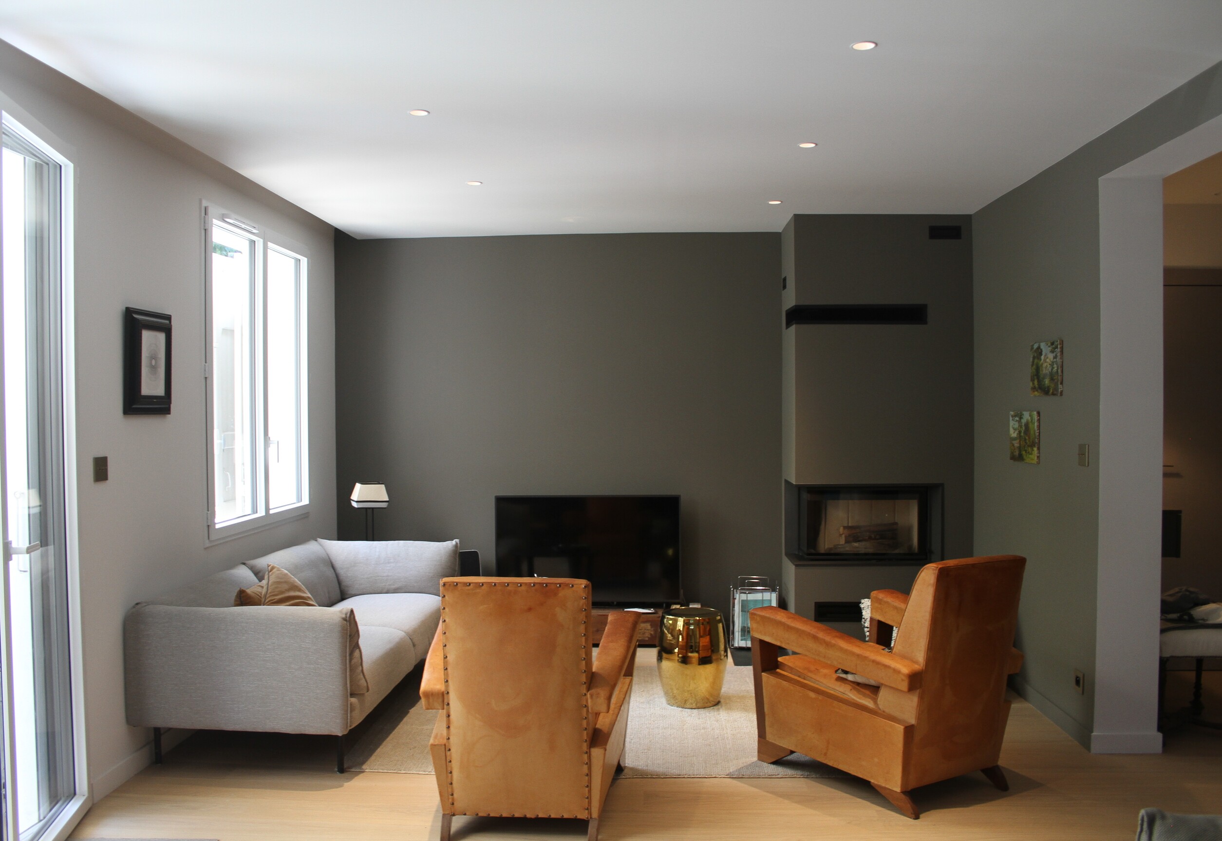 Salon moderne avec canapé gris, deux fauteuils marron, cheminée et télévision noire.