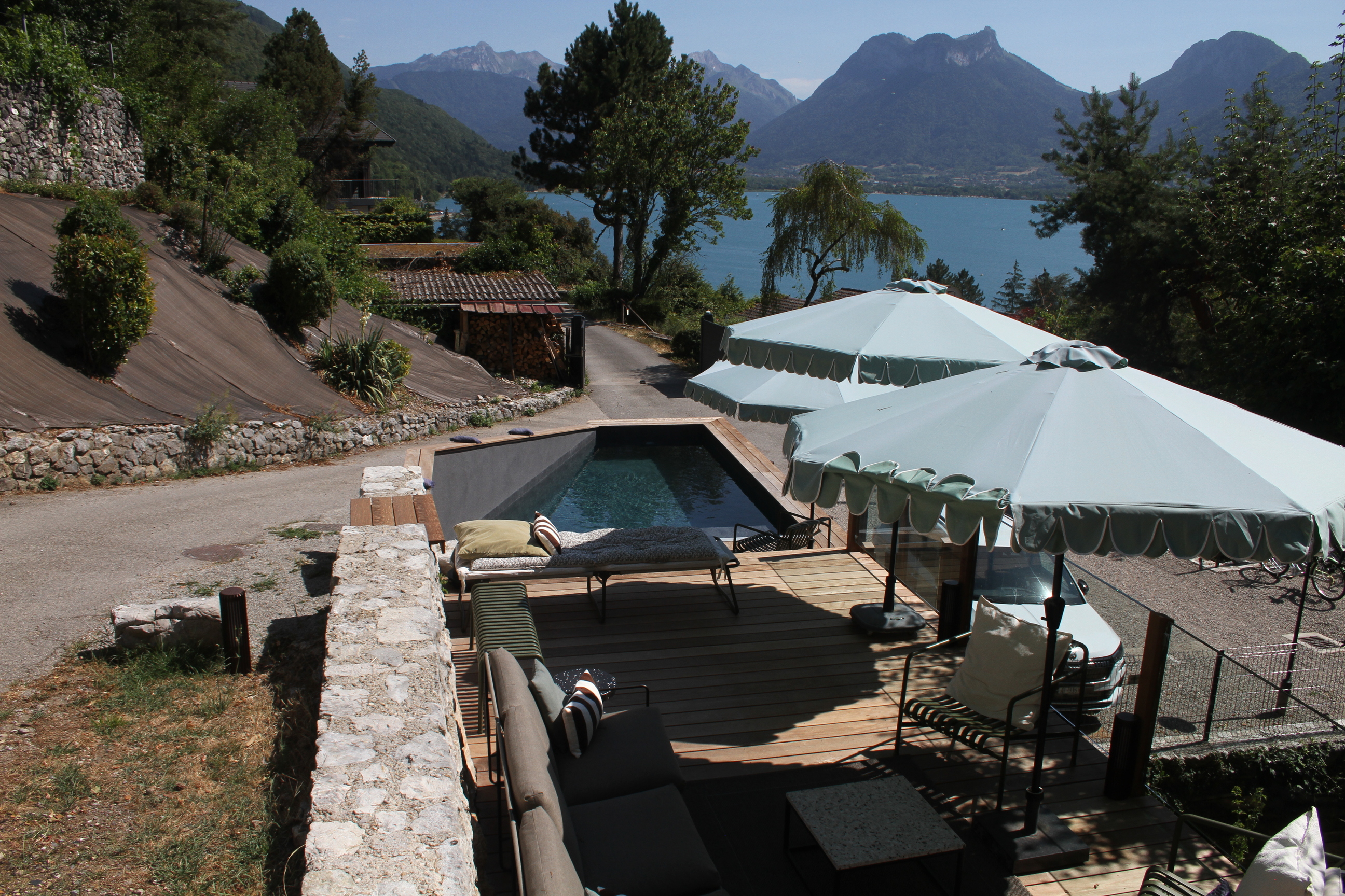 Terrasse en bois avec piscine, parasols bleus, canapés, vue panoramique sur un lac et montagnes en arrière-plan.