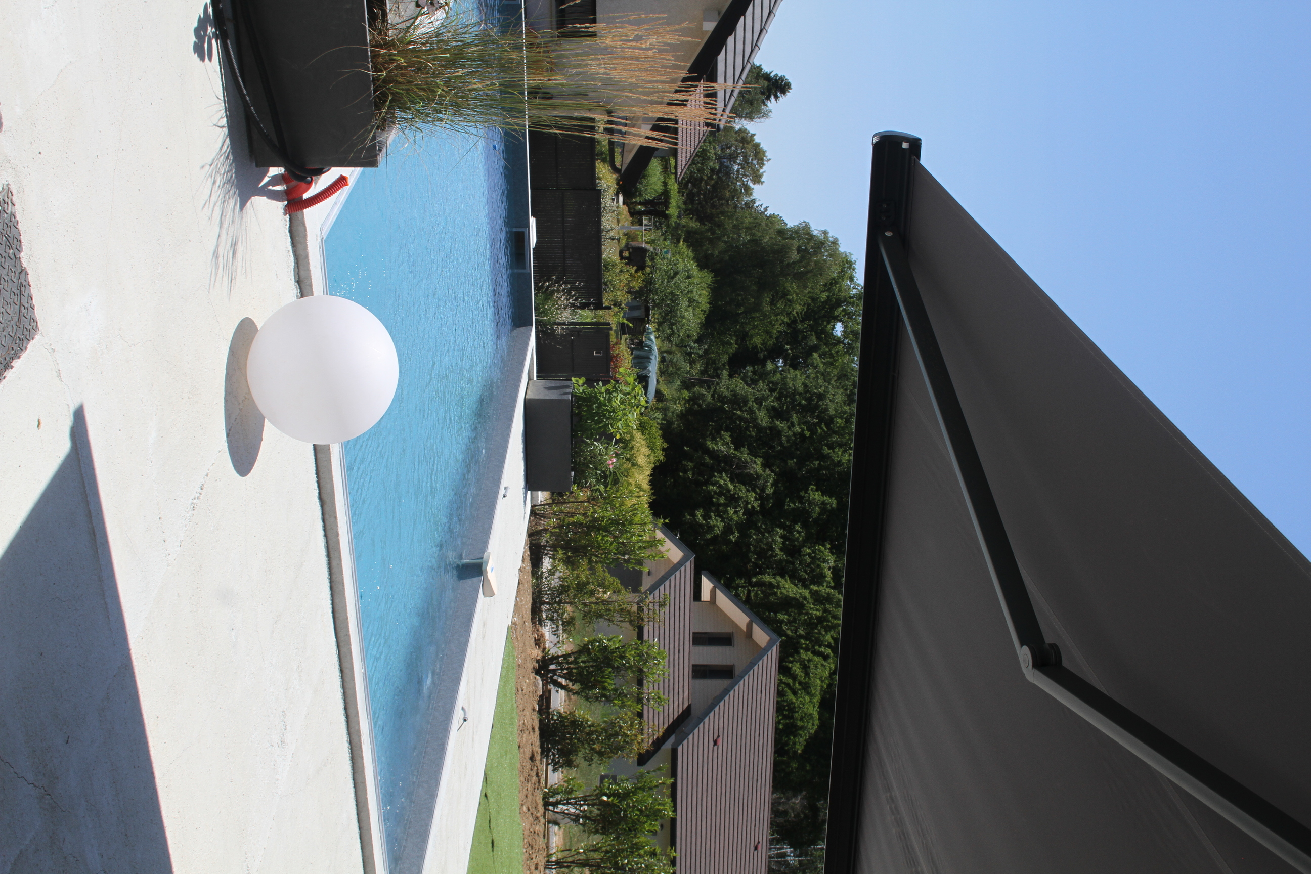 Piscine extérieure entourée d'une terrasse claire sous l'ombre d'un store banne.