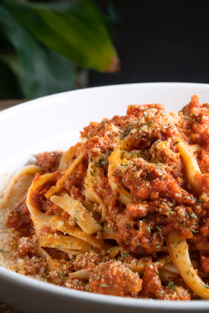 Close-up of tagliatelle pasta with rich meat Bolognese sauce topped with grated cheese and herbs.