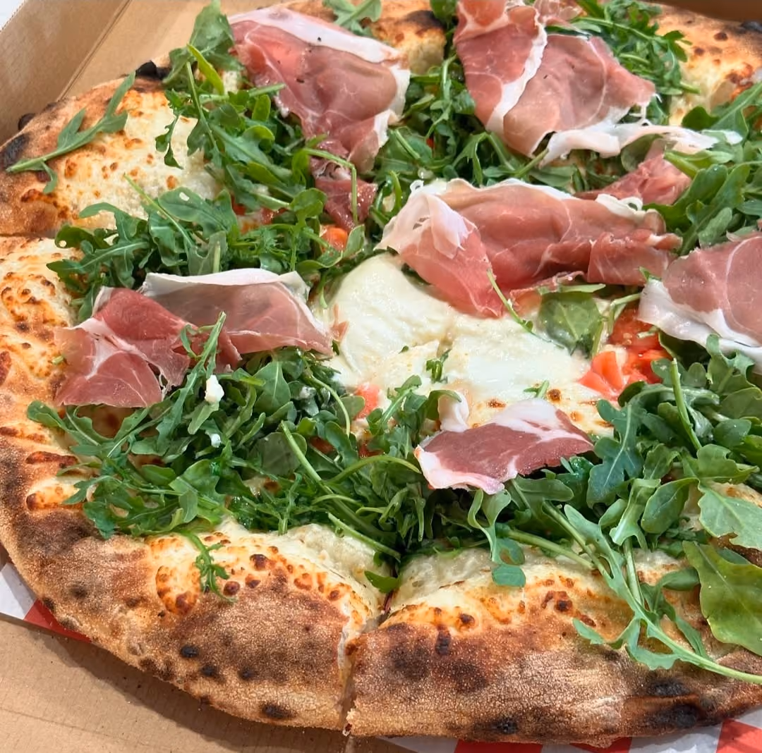 Pizza with melted cheese, arugula, and slices of prosciutto on a browned crust in a cardboard box.