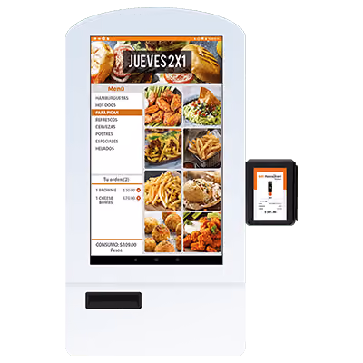 White self-service kiosk with a touchscreen menu showing food items like hamburgers, hot dogs, chicken wings, salads, fries, and desserts; the screen displays a 'Jueves 2x1' promo and an order summary with prices in pesos. Next to the screen is a smaller digital display for payment confirmation.