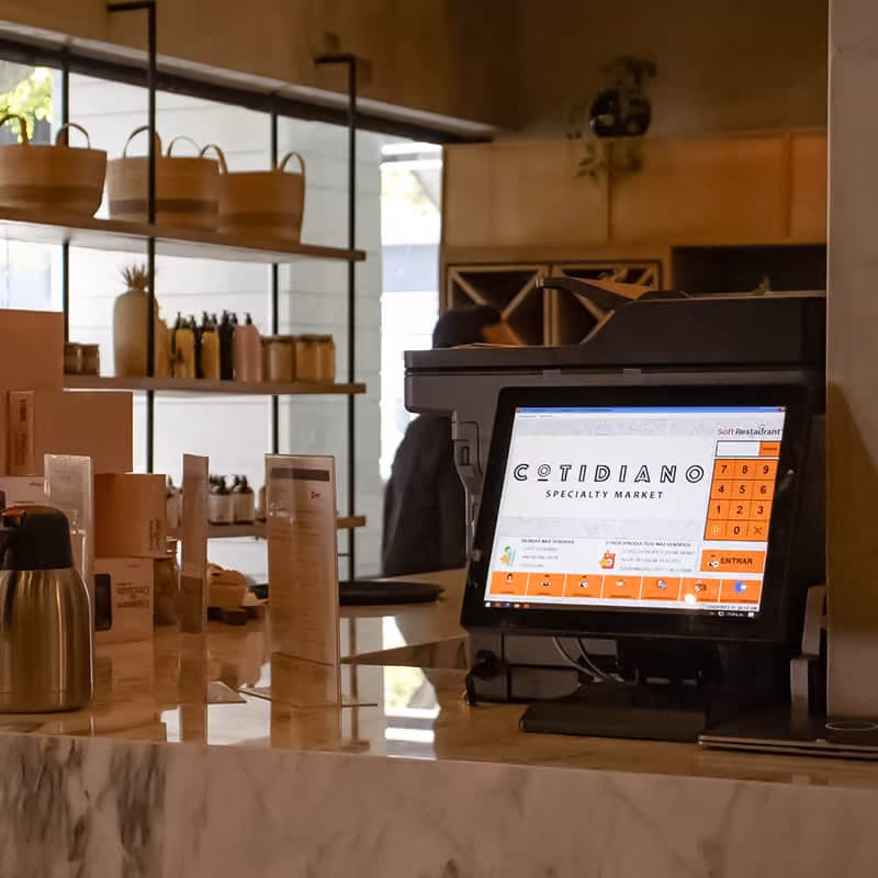 Retail or market checkout counter with a touchscreen point-of-sale system displaying the Cotidiano Specialty Market interface, surrounded by menus and containers on a marble countertop, with shelves and a person blurred in the background.