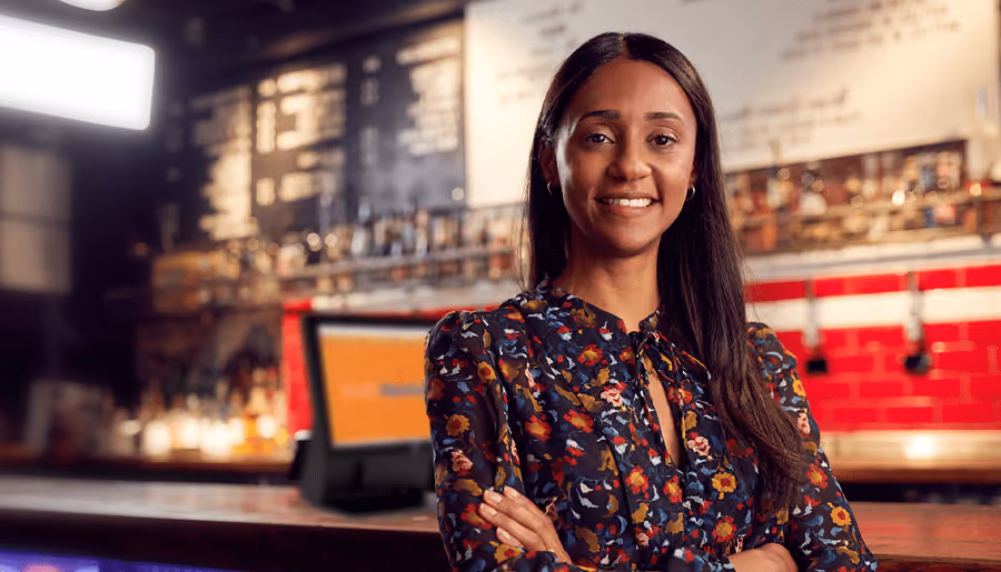 Smiling woman with long dark hair and a floral blouse standing with arms crossed in a warmly lit coffee shop with a menu board and counter in the background.