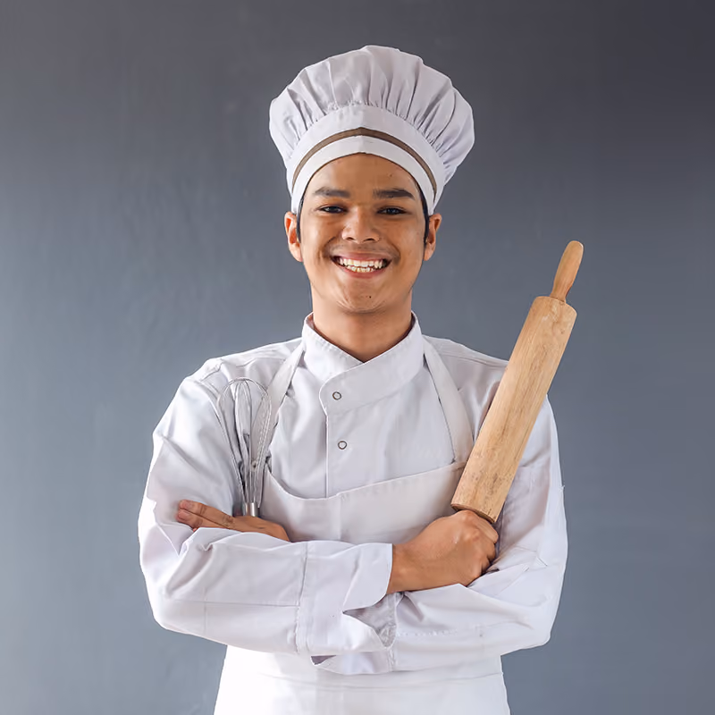 Smiling young chef in white uniform and hat holding a whisk and a wooden rolling pin with arms crossed against a gray background.