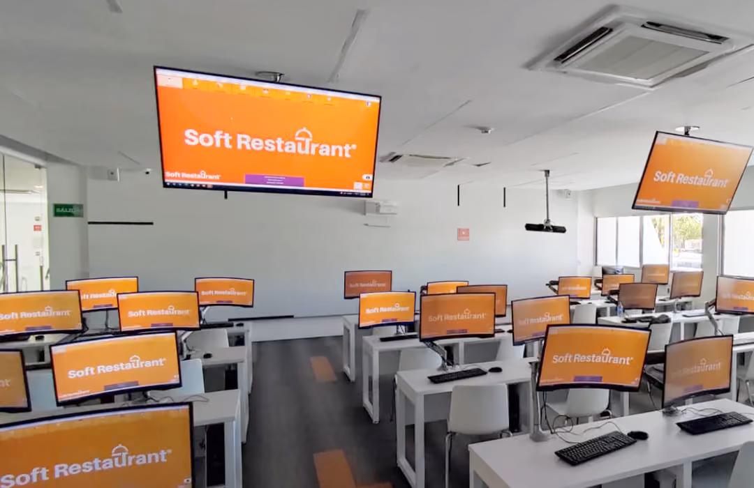 Classroom filled with computers on white desks, all displaying the Soft Restaurant logo on orange screens, with two large monitors hanging from the ceiling showing the same logo.