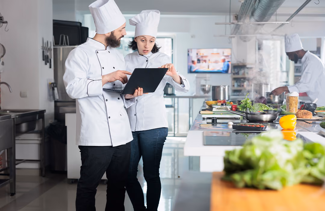 Two chefs in white uniforms and hats stand in a modern kitchen looking at a laptop; a third chef cooks in the background near a stovetop with various fresh ingredients on the counter.