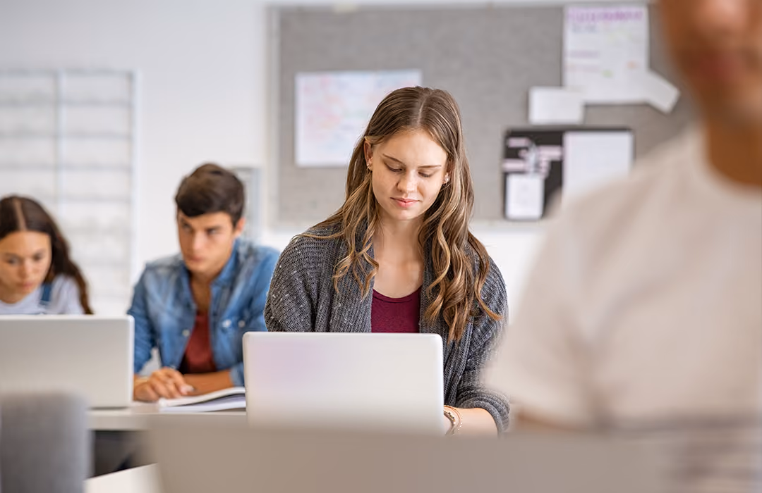 A young woman with long hair in a gray sweater sits at a desk using a laptop, with two other students focused on their work in the background inside a classroom.