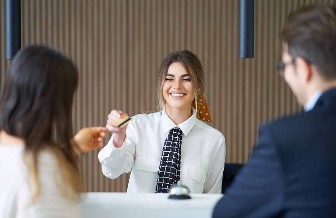 Smiling female receptionist in a white shirt with a patterned black tie handing a card to a customer across a white desk, with a man in a suit sitting nearby.