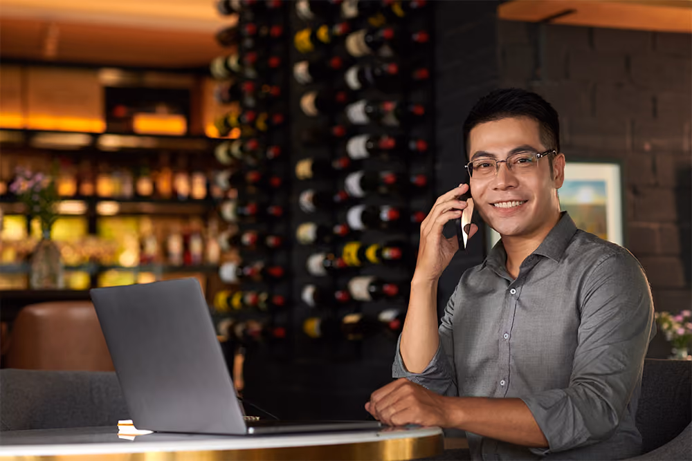 Smiling man wearing glasses and a gray shirt sits at a table with a laptop, talking on a smartphone in a warmly lit room with a wine bottle rack in the background.