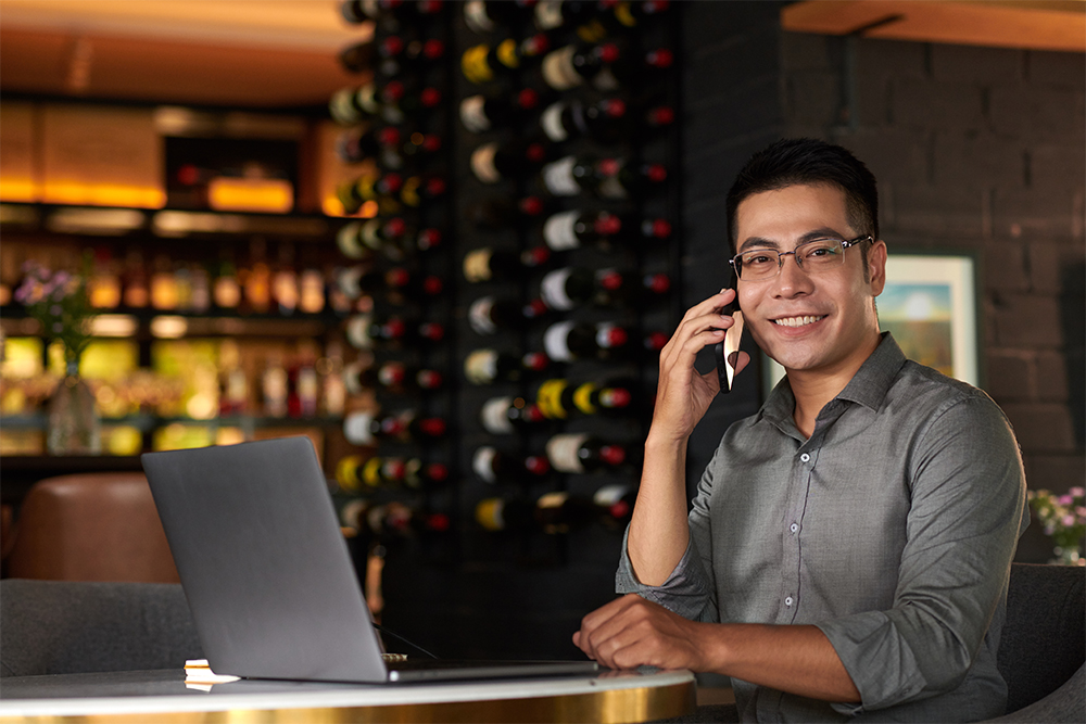 Smiling man wearing glasses and a gray shirt sits at a table with a laptop, talking on a smartphone in a warmly lit room with a wine bottle rack in the background.