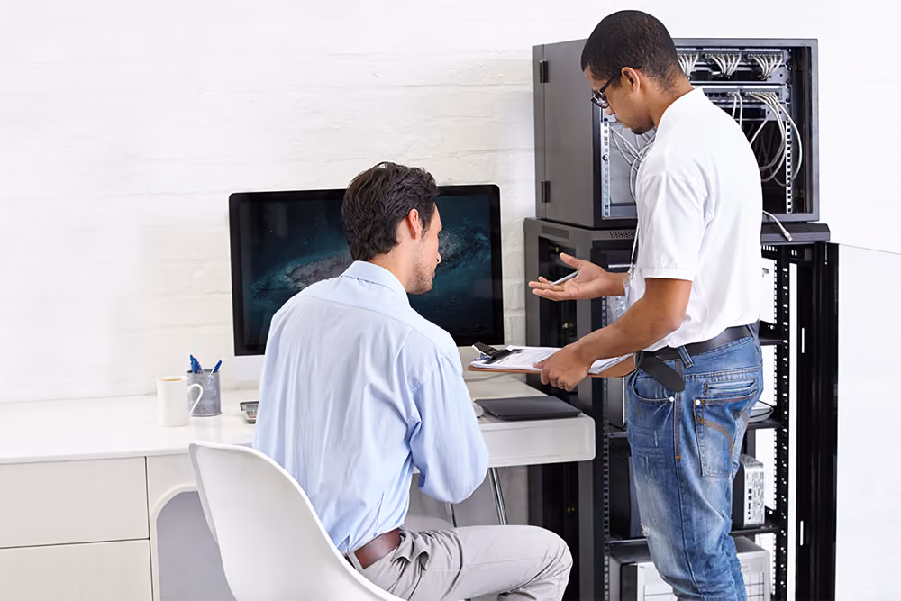 Two men in an office setting working with computer equipment; one man in a white shirt and jeans holds a clipboard and gestures while standing near a server rack, the other man in a light blue shirt sits at a desk with a large monitor, listening.