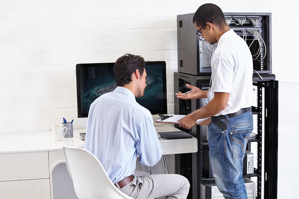 Two men in an office setting working with computer equipment; one man in a white shirt and jeans holds a clipboard and gestures while standing near a server rack, the other man in a light blue shirt sits at a desk with a large monitor, listening.