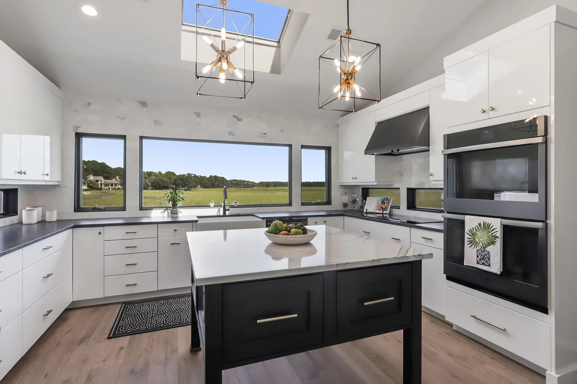 Modern kitchen with white cabinets, black countertops, a central island with white marble top and black base, two pendant lights, large windows showing a green landscape, built-in double ovens, and a farmhouse sink under the wide window.