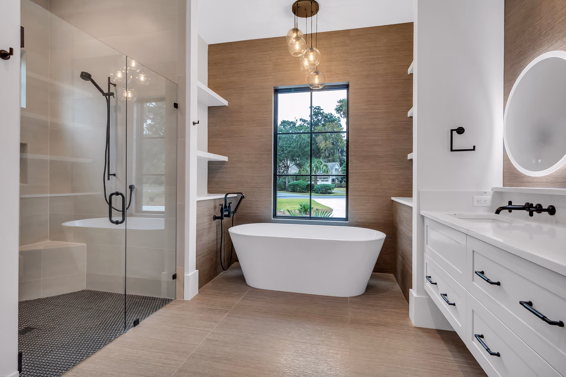 Modern bathroom with a white freestanding bathtub centered beneath a large window, beige tile walls and floor, glass-enclosed shower with black fixtures on the left, and white vanity with black handles and a round illuminated mirror on the right.