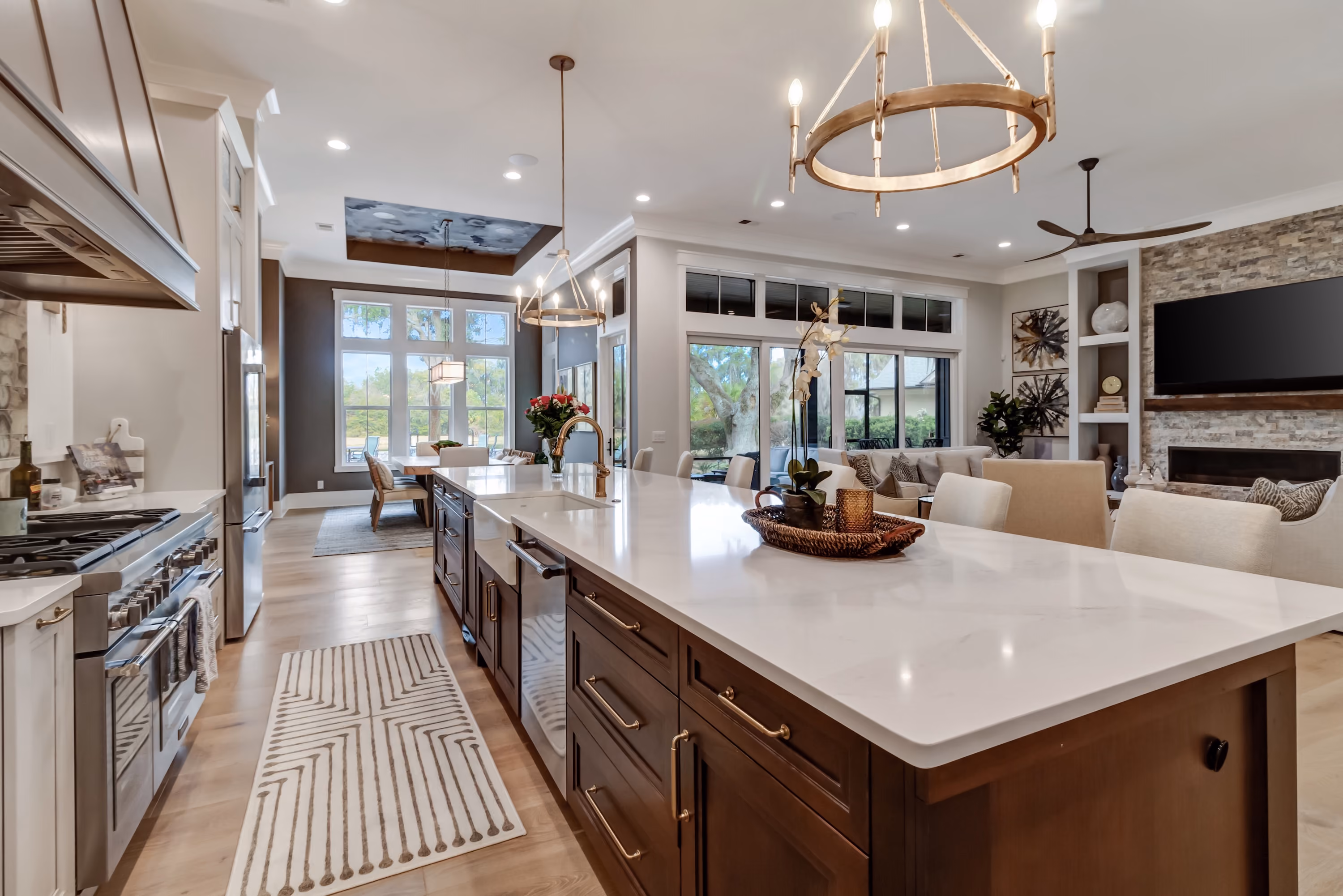Modern open kitchen and living room with a large white marble island featuring dark wood cabinets, stainless steel appliances, and decorative tray with orchid. The living area has beige chairs and a stone accent wall with a mounted flat screen TV and built-in shelves, adjacent to sliding glass doors with a view of greenery outside. Pendant and chandelier lighting illuminate the space, and wood flooring runs throughout.