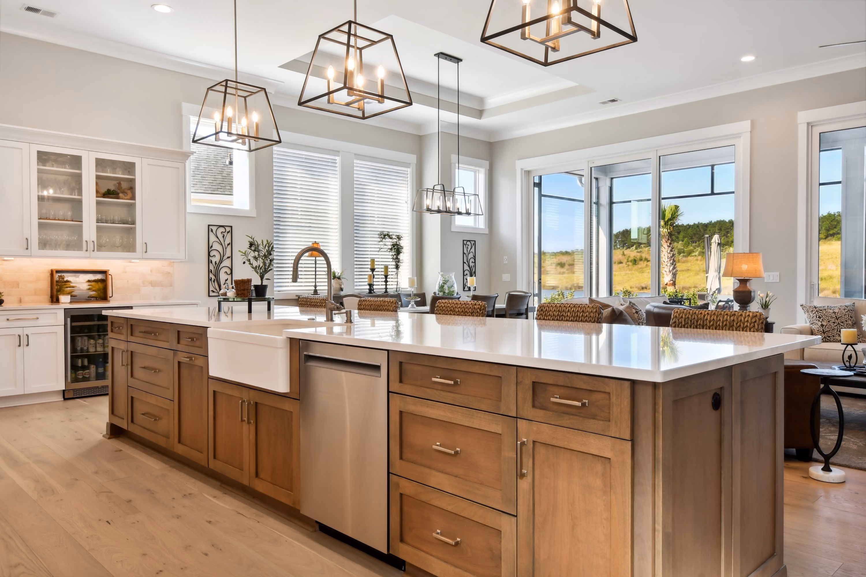 Bright modern kitchen with a large wooden island featuring a white countertop, farmhouse sink, and stainless steel dishwasher, surrounded by wicker bar stools and illuminated by three pendant lights, with windows and sliding glass doors in the background showing an outdoor view.