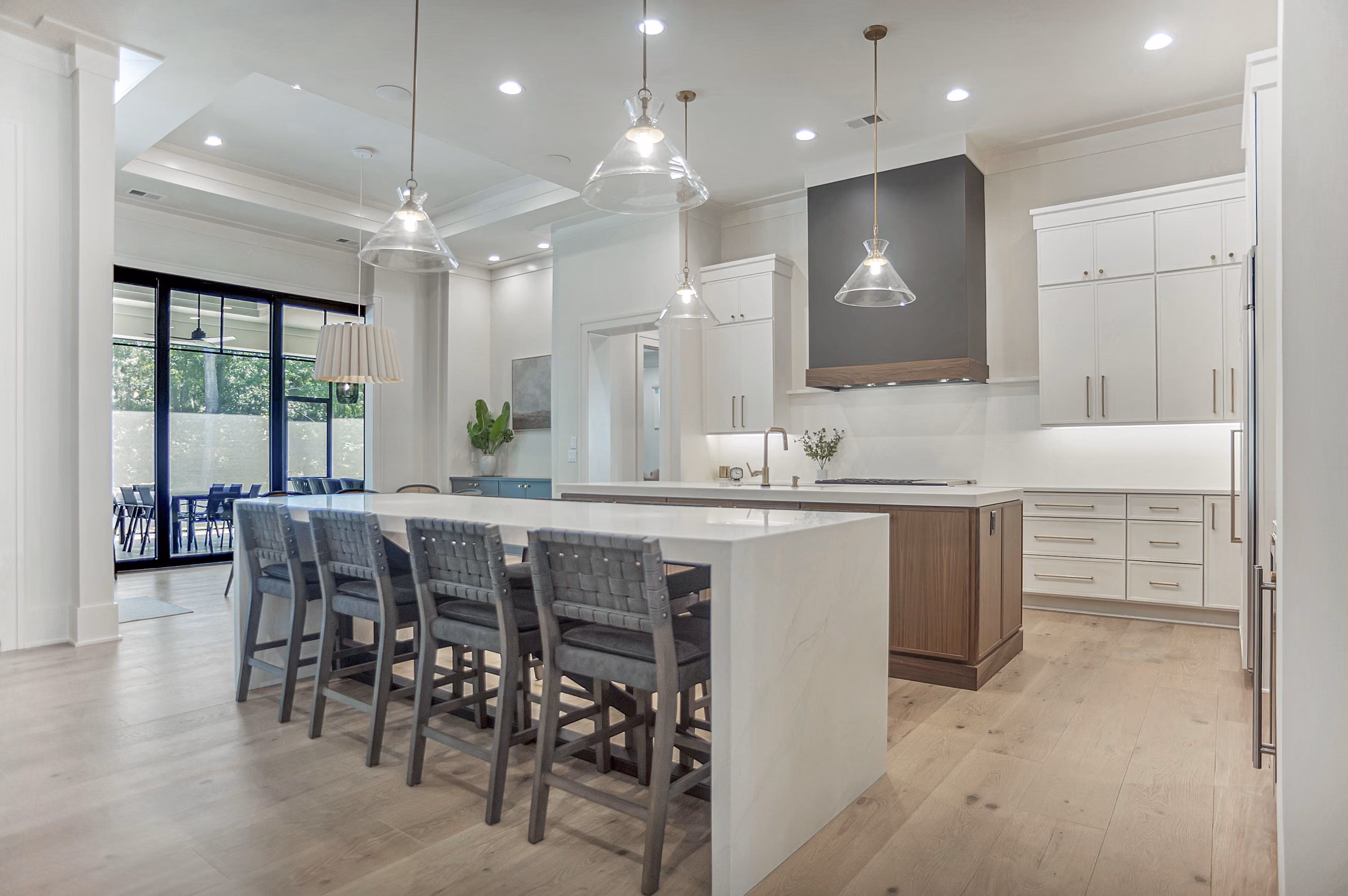 Modern kitchen with a large white island featuring six gray cushioned bar stools, pendant lights hanging above, white cabinetry with gold handles, wood flooring, and a dark gray range hood.