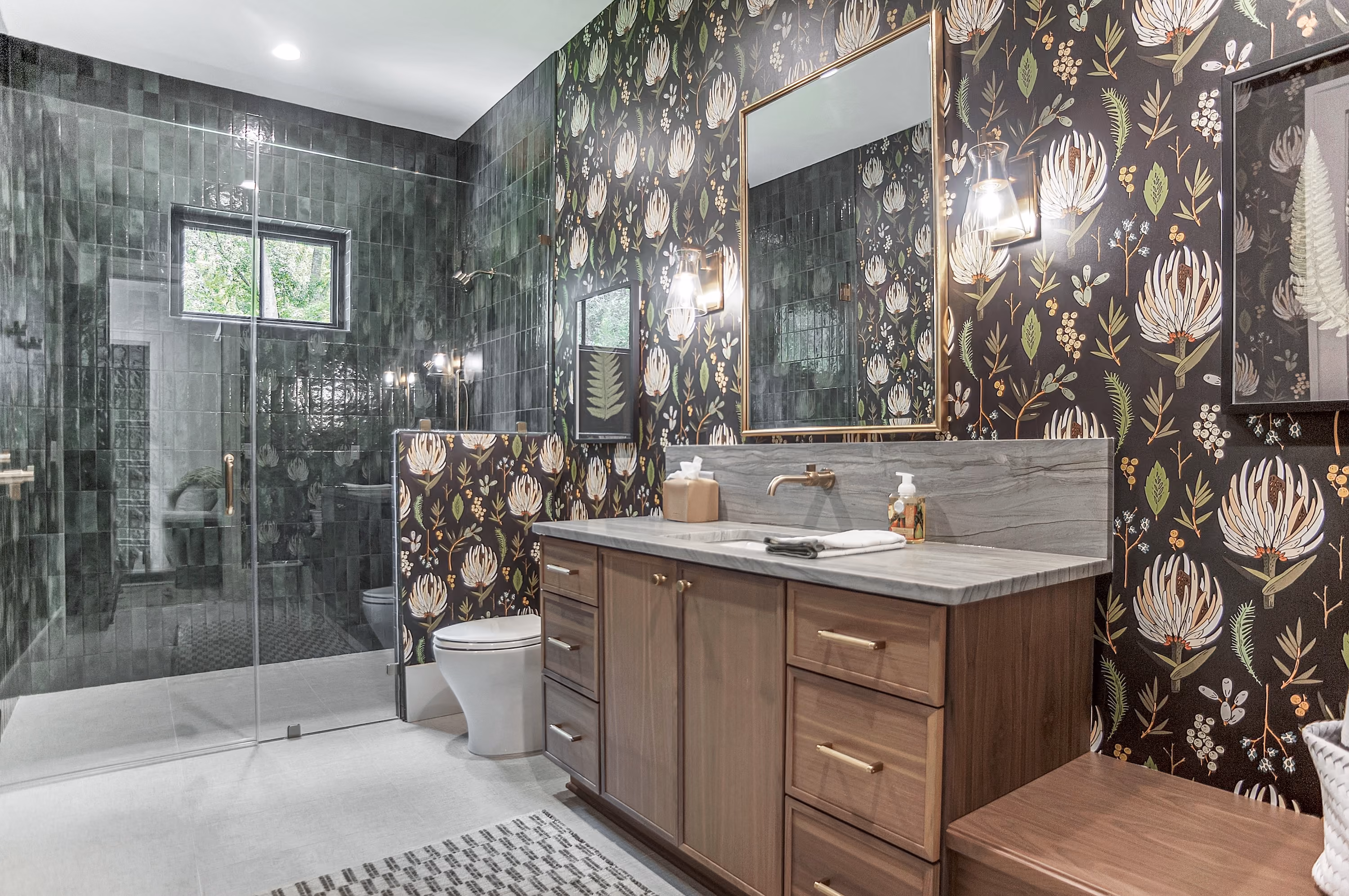 Modern bathroom with floral wallpaper featuring large white flowers and greenery, a wooden vanity with marble countertop and brass fixtures, two framed botanical prints, and a glass-enclosed shower with dark green tiles and a small window.