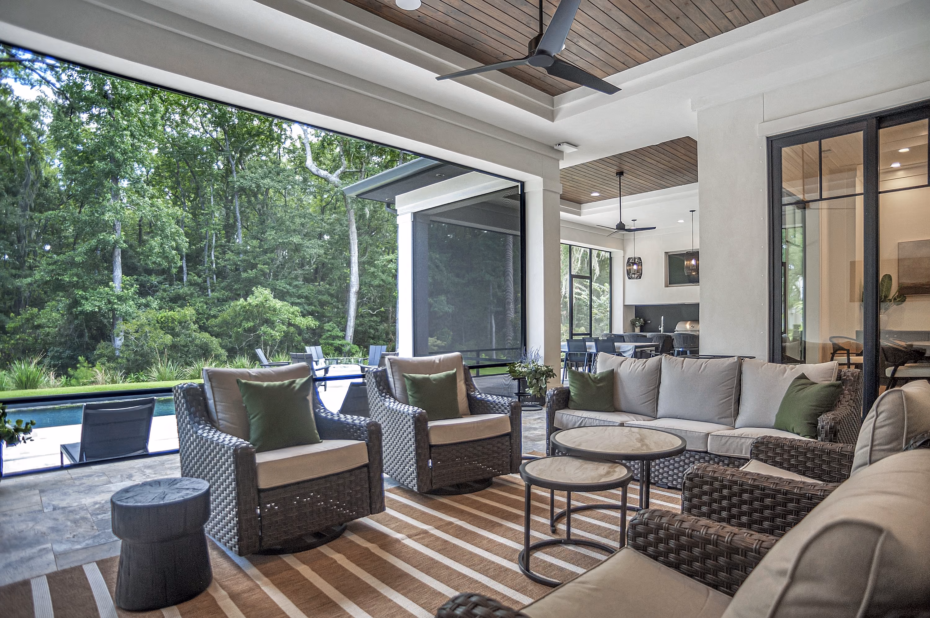 Covered outdoor patio with grey woven furniture including sofas and armchairs with beige cushions and green pillows, two round nested coffee tables, a side table, ceiling fans, and a striped area rug, overlooking a swimming pool and lush green trees.