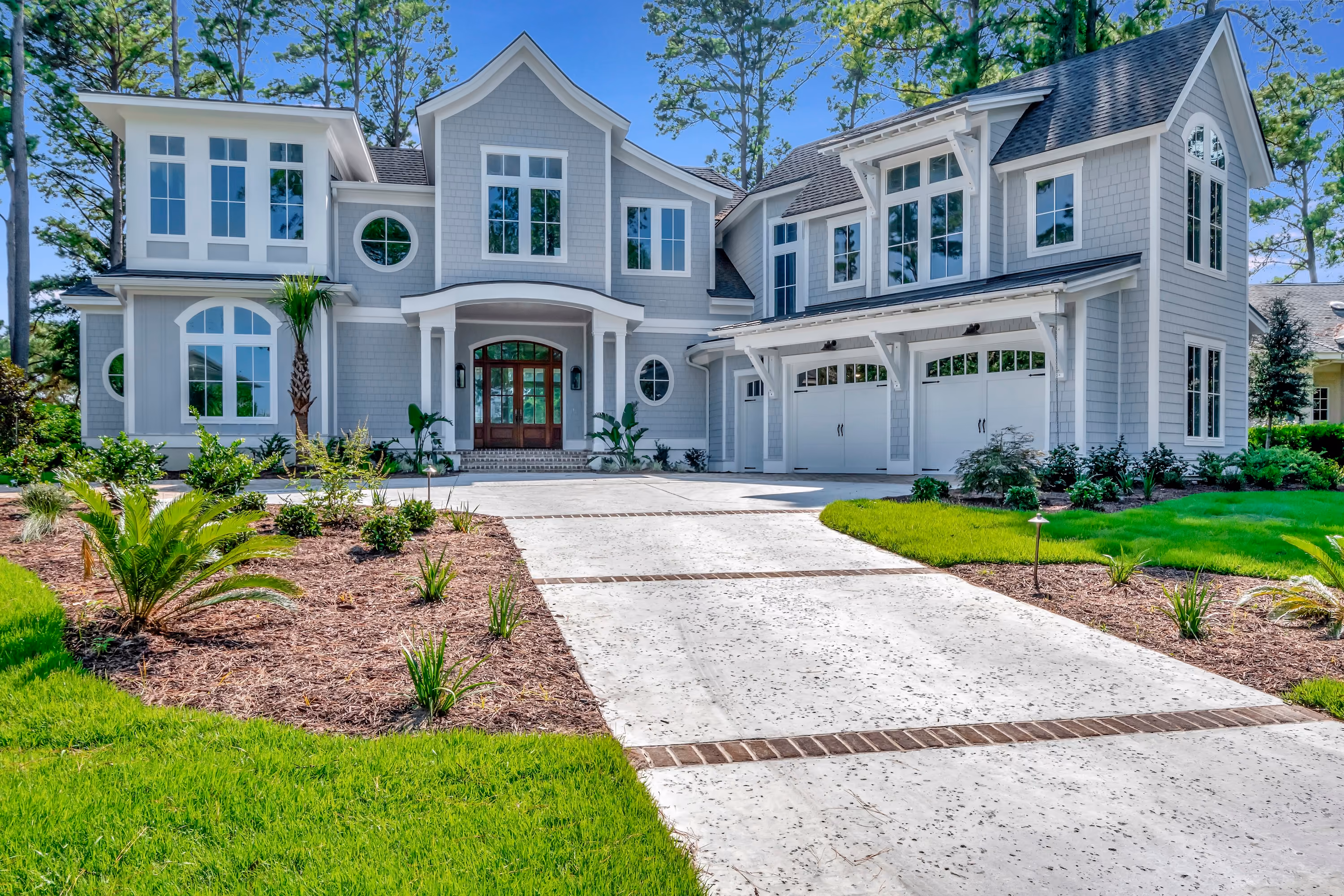 Large two-story gray house with multiple windows, a curved concrete driveway with brick accents, a three-car garage, landscaped garden beds with small plants and palm trees, and a front porch with a wooden door under a gabled roof, set against clear blue sky and tall trees.