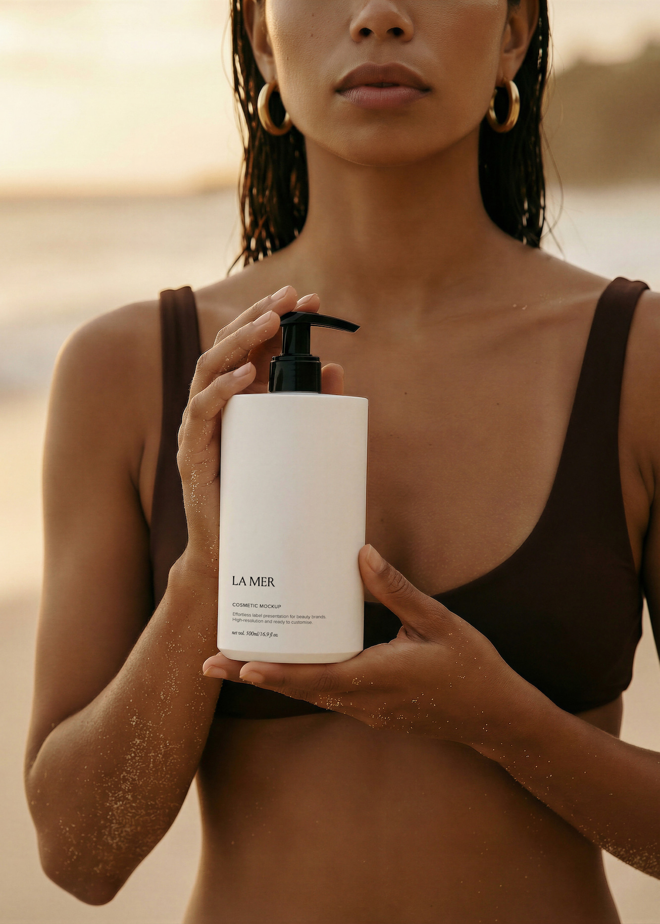 A woman with wet hair and gold hoop earrings in a brown bikini top holds a white cosmetic pump bottle mockup labeled 'LA MER' on a sandy beach.