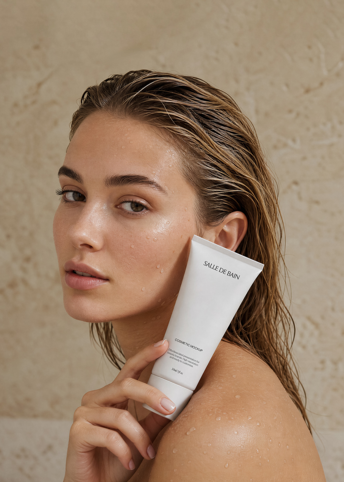 Close-up of a woman with wet skin and hair holding a white cosmetic tube mockup labeled 'Salle de Bain' near her cheek against a neutral textured background.