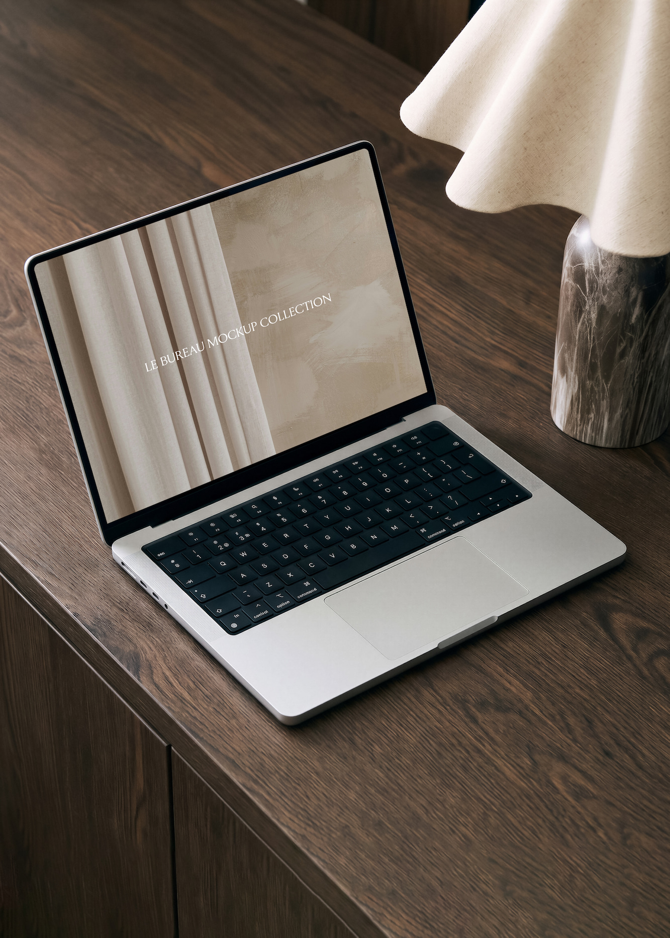Open MacBook mockup on a wooden desk with a screen displaying beige curtains and the text 'LE BUREAU MOCKUP COLLECTION'; a marble-based lamp with a cream-colored shade is on the right side of the desk.