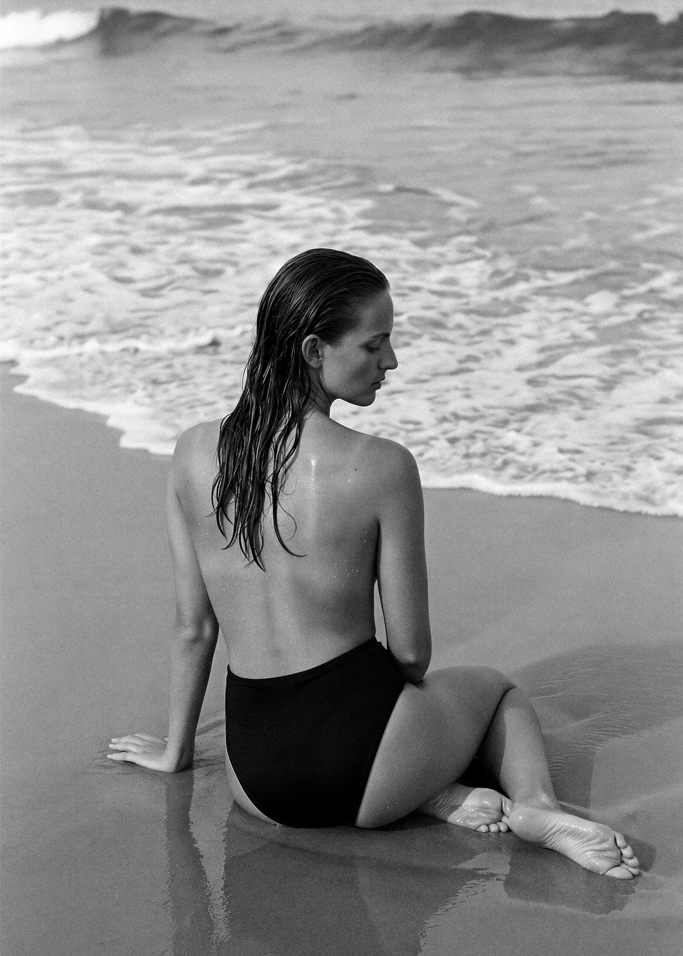 Black and white photo of a woman sitting on wet sand at the beach, viewed from behind, with wet hair and wearing high-waisted black swimwear as waves gently approach the shore.