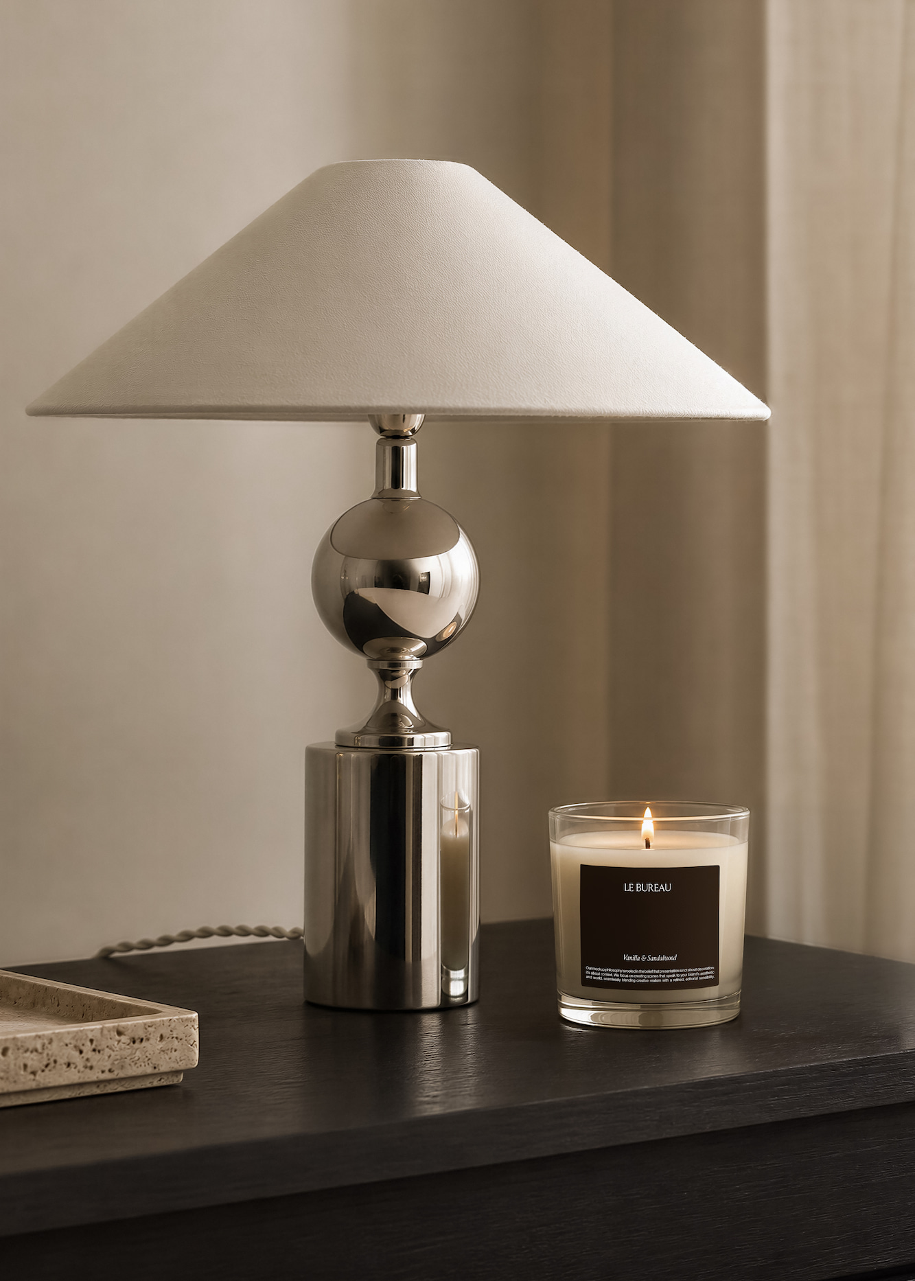 A modern table lamp with a metallic base and a cream-colored conical lampshade sits on a dark wooden surface next to a lit scented candle in a clear glass container labeled 'LE BUREAU Vanilla & Sandalwood.'
