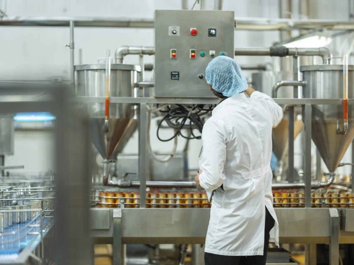 Food production line worker in a hairnet and white coat checking a stainless steel filling line