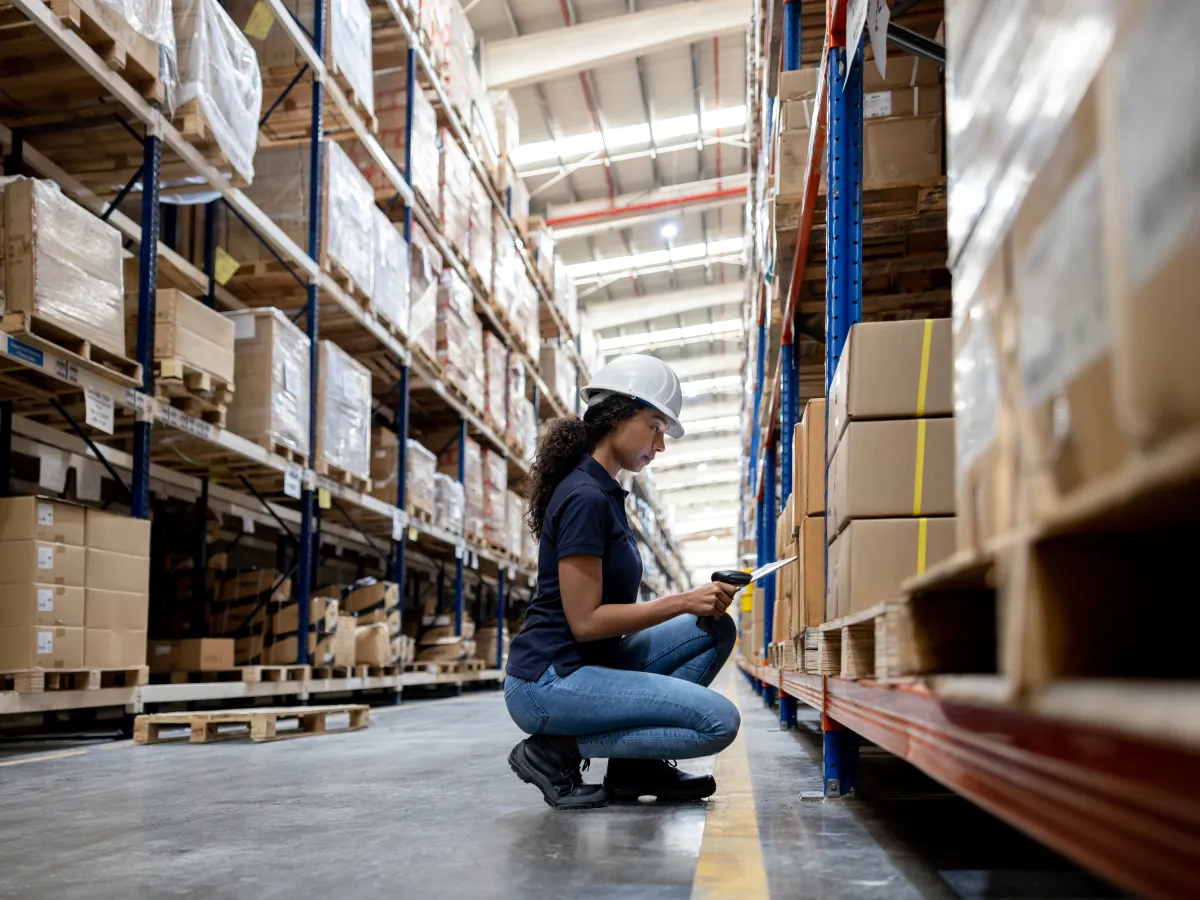 Warehouse worker in a hard hat crouching to scan a pallet label on a low rack with a handheld scanner