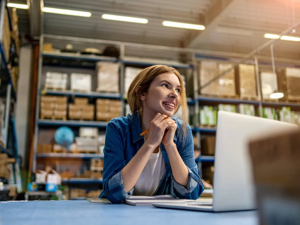 Warehouse employee in a denim shirt sitting at a laptop in front of stocked shelving, chin resting on her hands