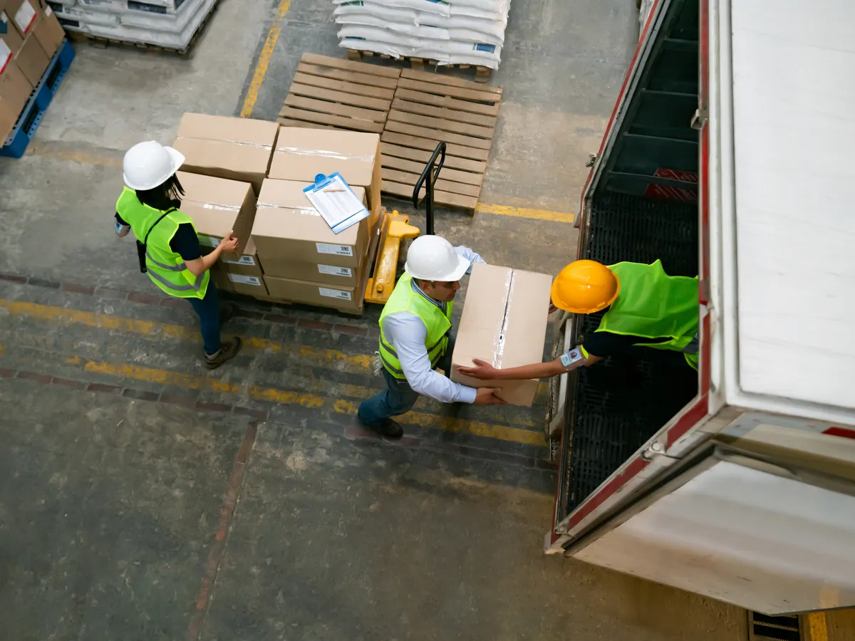 Overhead view of three workers in hard hats moving cartons between a trailer and a pallet jack, with a clipboard of paperwork on the stack