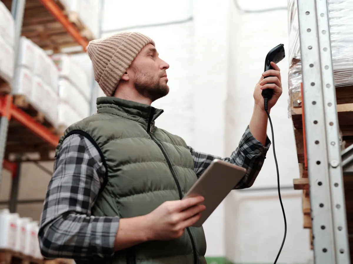 Warehouse worker in a knit cap and vest scanning a pallet on a rack with a handheld scanner, tablet in his other hand