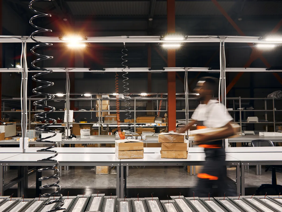 Packing benches in a fulfillment center behind a roller conveyor, a worker in a white shirt carrying boxes past in motion blur